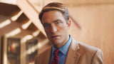 A man in a tan suit, light blue dress shirt, and red patterned tie, shown from the chest up in a warm-toned indoor setting.