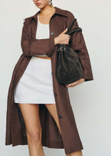 A woman models a long brown trench coat over a white crop top and white mini skirt, holding a dark brown suede shoulder bag against a plain light gray studio background.