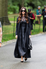 Woman in a black dress and coat walking outdoors with blurred background
