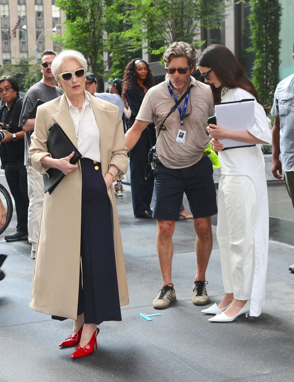 Woman in a beige coat and red shoes walking on a city street with other people around.