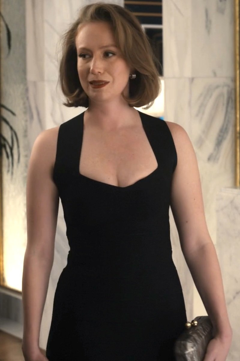 A woman with short brown hair wearing a sleeveless black dress with a deep scoop neckline, standing in an indoor setting with marble walls.