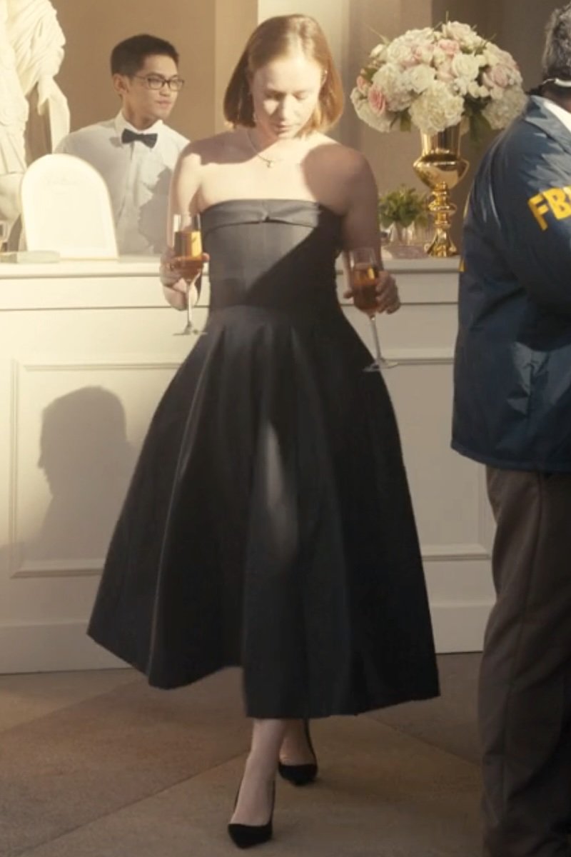 A woman wearing a strapless black midi dress with a structured bodice and full skirt, holding two champagne glasses in a formal indoor setting.