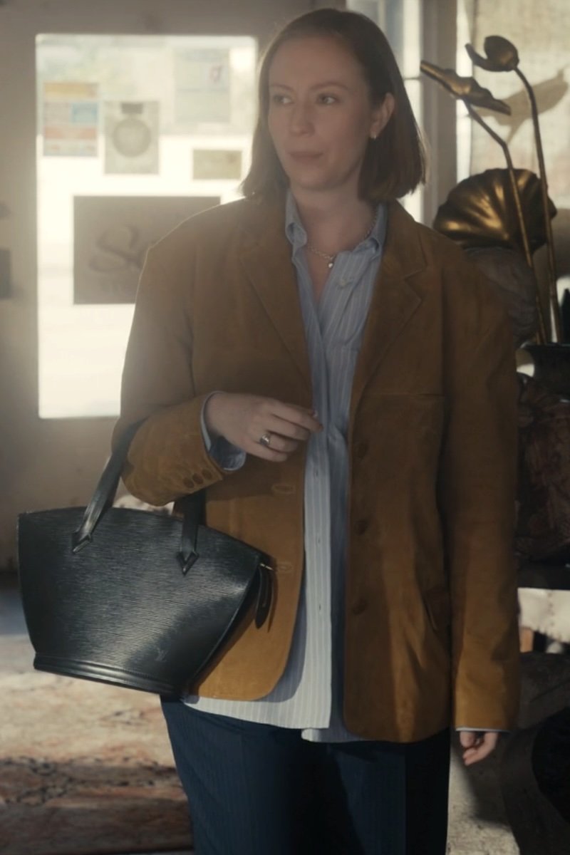 A woman wearing a brown suede blazer over a light blue striped shirt, holding a black textured leather handbag in a softly lit indoor setting.