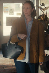 A woman wearing a brown suede blazer over a light blue striped shirt, holding a black textured leather handbag in a softly lit indoor setting.