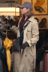 A woman wearing a beige trench coat, gray cap, dark scarf, and brown gloves stands in a shop setting.