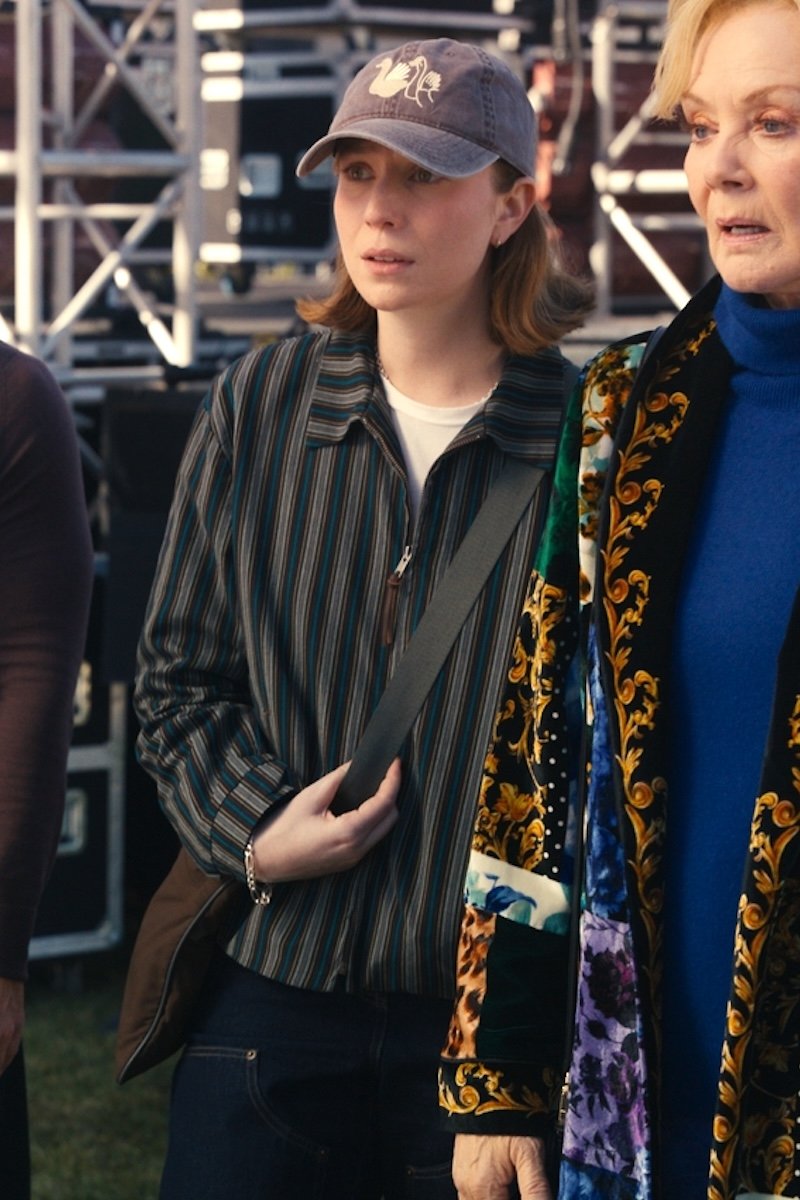 A woman wearing a dark striped button-up shirt, dark jeans, a gray baseball cap, and a brown shoulder bag stands outdoors next to another person in a patterned coat.
