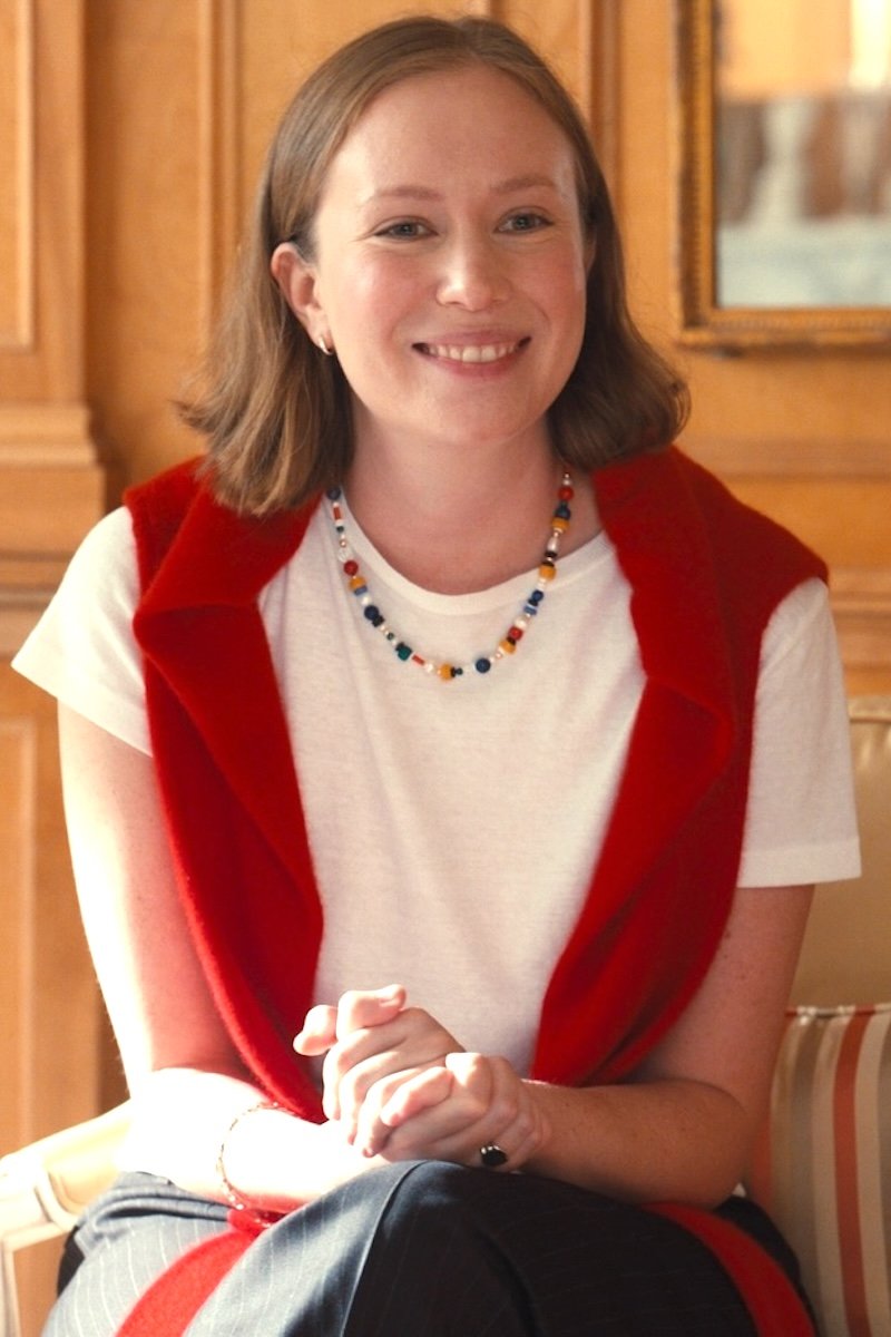 Woman wearing a red vest over a white shirt with a colorful necklace, sitting indoors.
