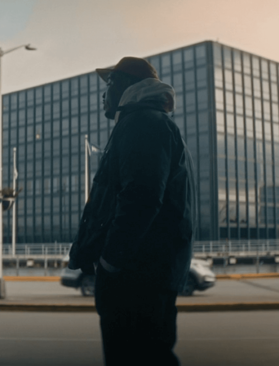 A person in a dark winter jacket and cap stands in profile on a city street in front of a large modern glass building.