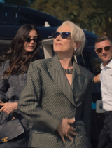 Woman in a green pinstripe suit with sunglasses, standing outdoors with blurred people in the background.
