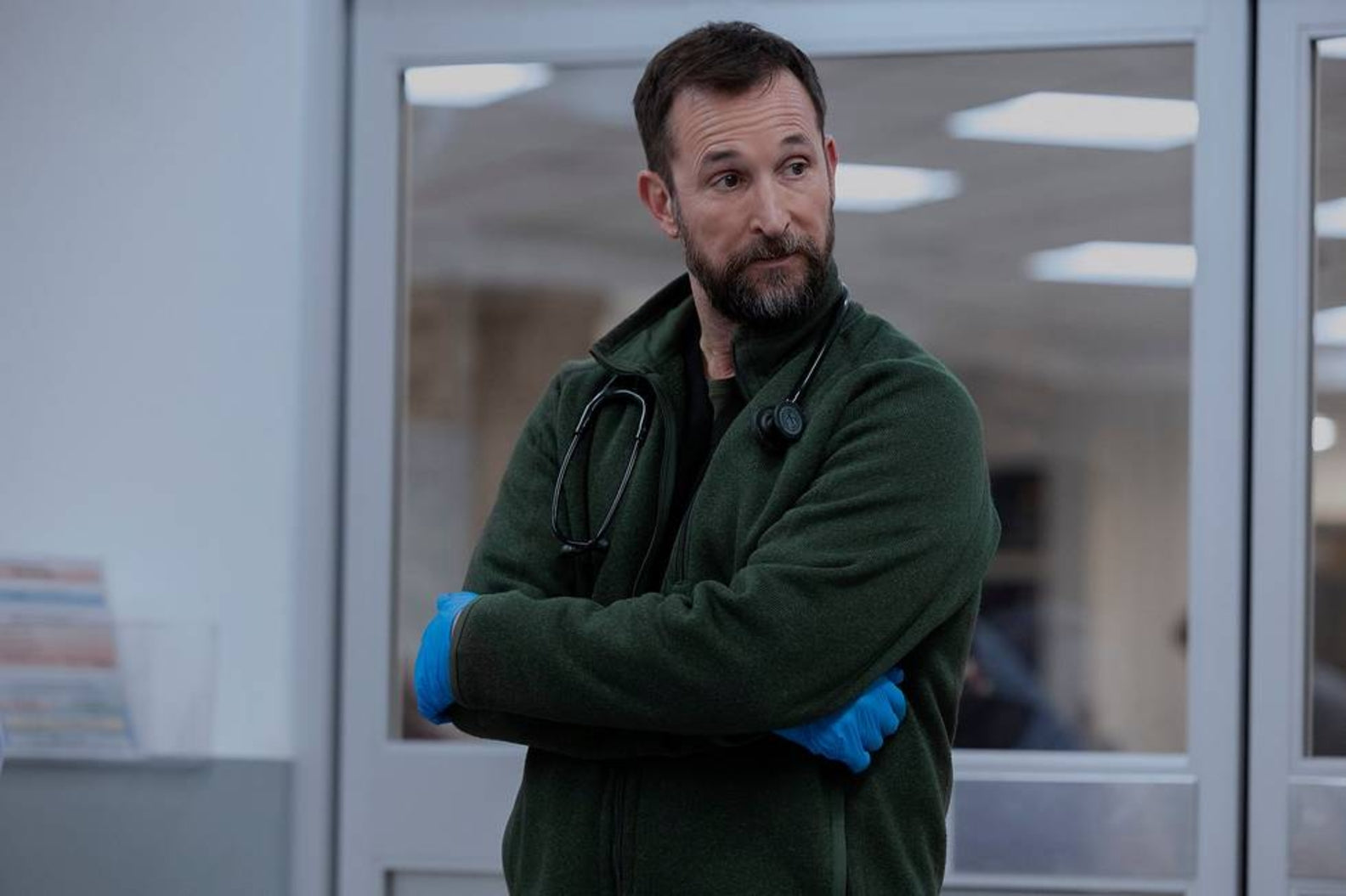 Man in a green coat with stethoscopes around his neck, standing in a room with glass doors.