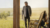 Man standing in a field with a wooden chair and plaid blanket nearby.