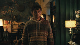 A man wearing a brown and beige patterned knit hoodie stands in a dimly lit room with warm lamps and dark green walls in the background.