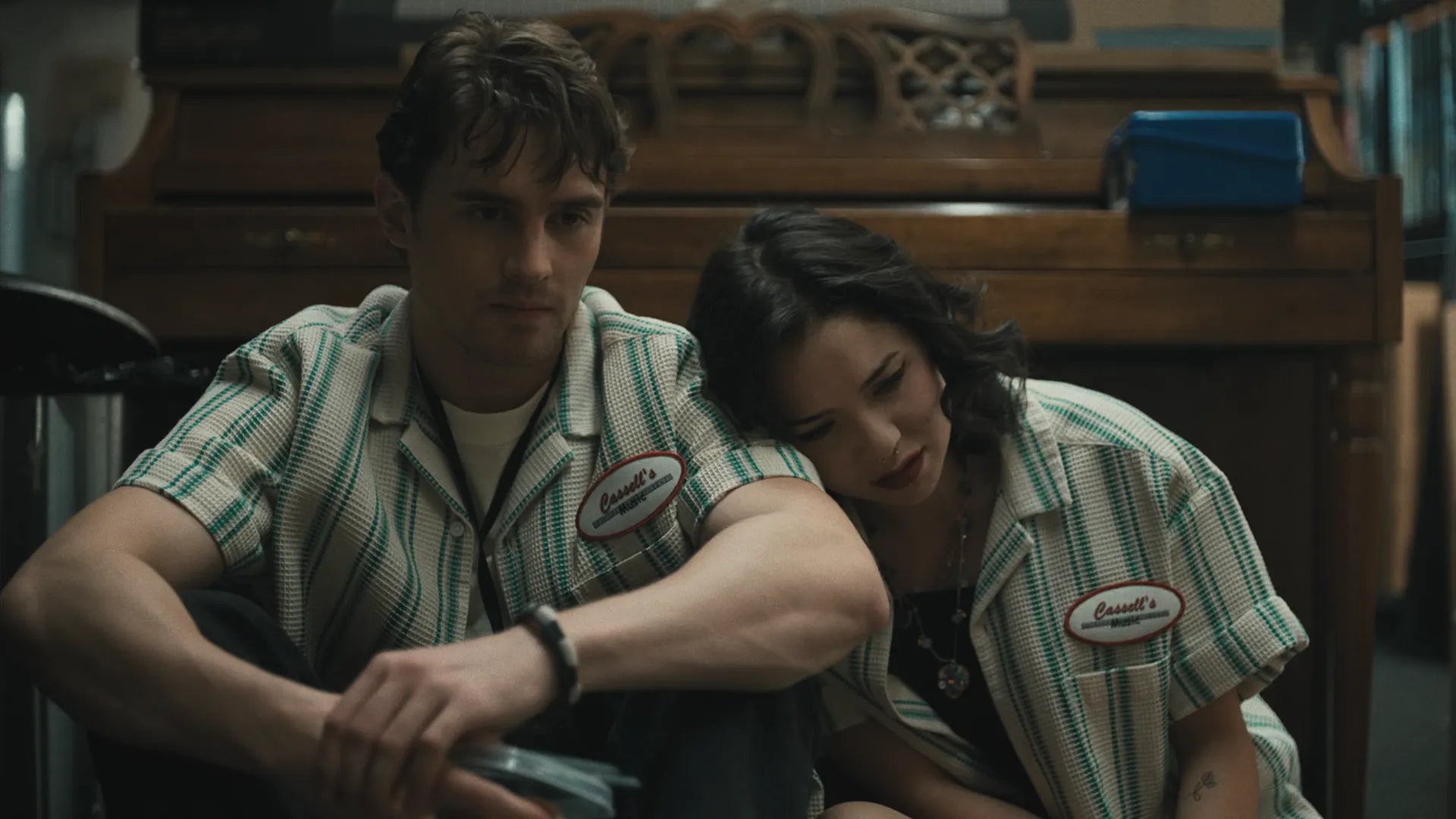 A man and a woman wearing matching green and white striped diner-style shirts with 'Bessie's' patches, seated indoors in a dimly lit restaurant or cafe setting.