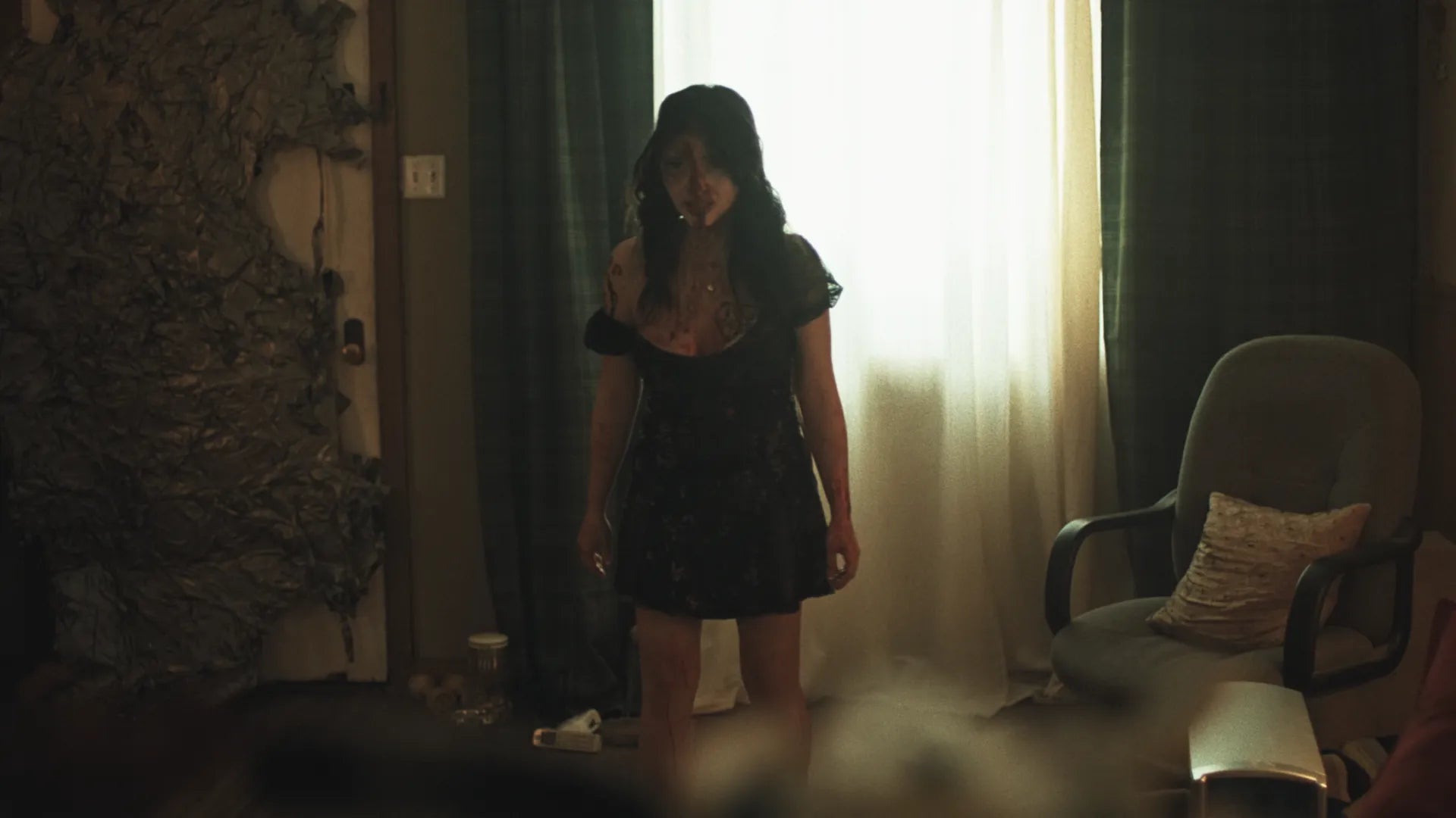 A woman in a short black dress stands in a dimly lit room near a bright window with curtains, with a chair and textured wall visible in the background.