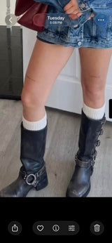 A person wearing black knee-high leather boots with silver buckle details, white socks, and denim shorts in an indoor setting.