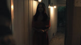 A woman with long dark hair stands in a dimly lit hallway at night, wearing a dark red dress. Warm light glows from wall-mounted bulbs on both sides of the corridor.