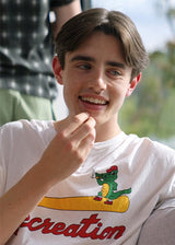 A split-screen photo showing a young man wearing a white short-sleeve henley T-shirt with a red 'Recreation' script and a green cartoon alligator graphic. The left side is a casual outdoor close-up, and the right side is a clean studio-style full-body shot against a light neutral background.