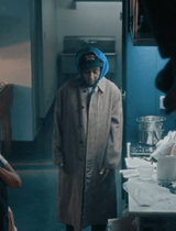 A person wearing a long taupe trench coat, a blue hood, and a black baseball cap stands in a dimly lit kitchen or utility room.