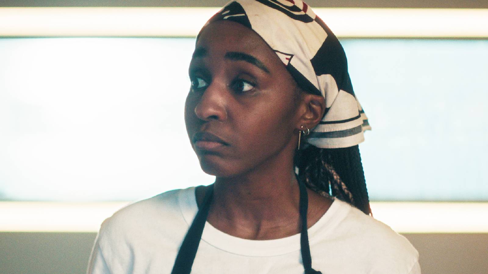 A woman wearing a black and white patterned headscarf and a white t-shirt with a black apron strap, looking off to the side against a bright, softly lit background.