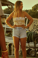 Woman in a white lace top and denim shorts standing outdoors with trees and a sign in the background.