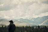 Person wearing a hat and coat standing in front of a mountain range with trees in the foreground.