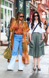 Two women walking on a city street, one in an orange dress and blue pants, the other in a white shirt and green skirt.